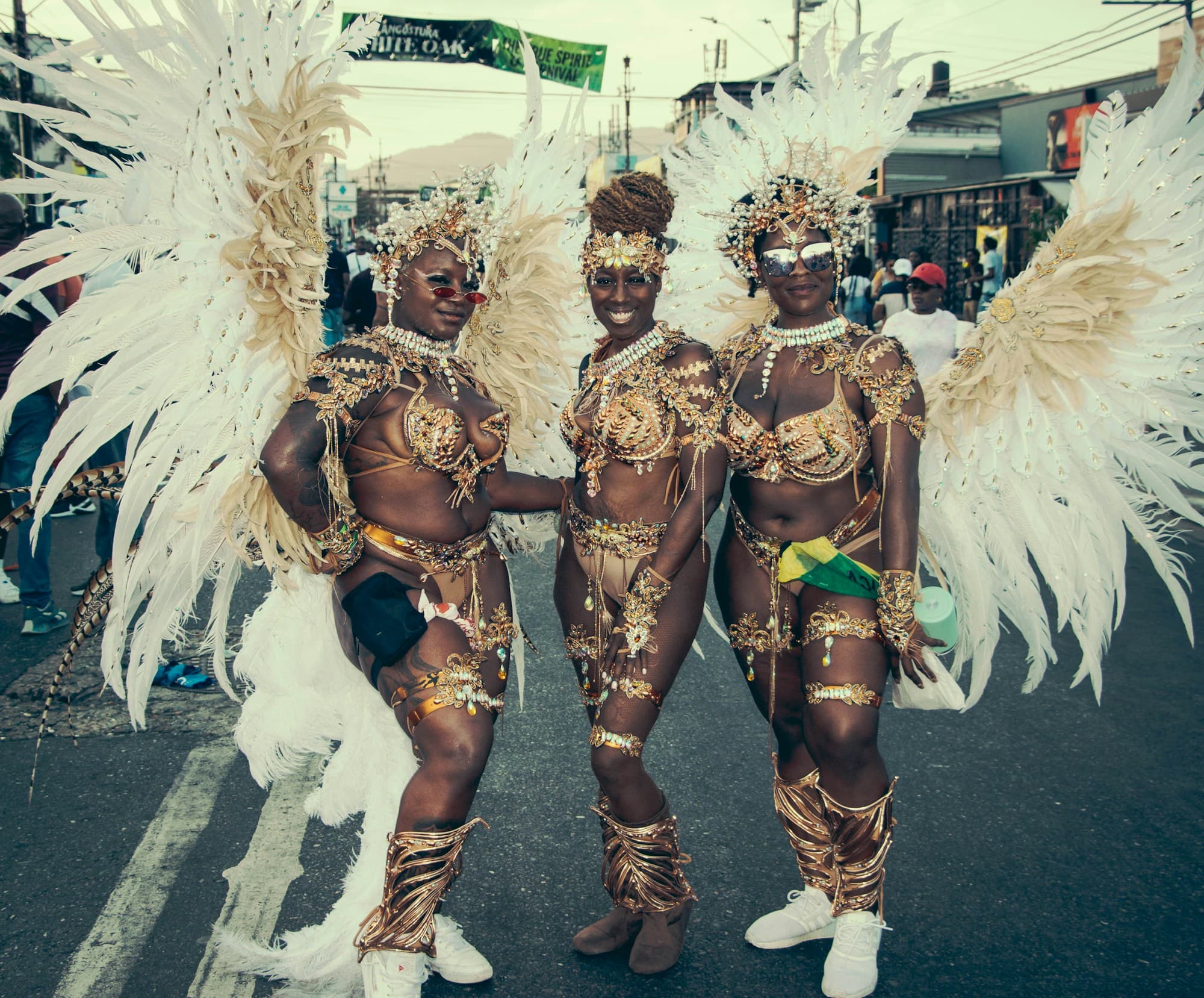 Three masqueraders in gold beaded bikinis, gold leg pieces and large white feathered backpacks stand together on a city street, with spectators and shopfronts behind them