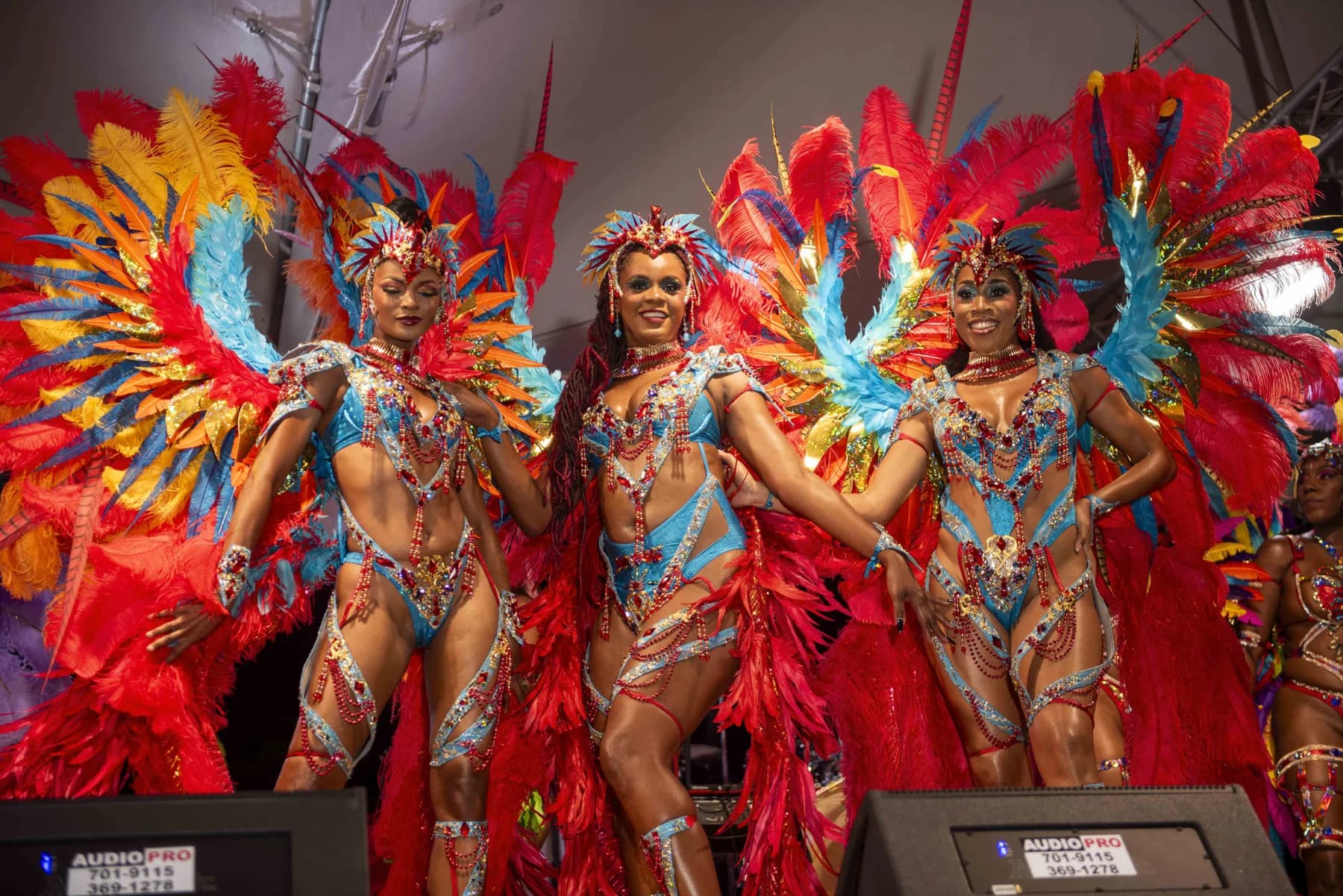 Three Showtime masqueraders in red, orange and blue feathered Take Off costumes on stage at the Carnival 2027 band launch