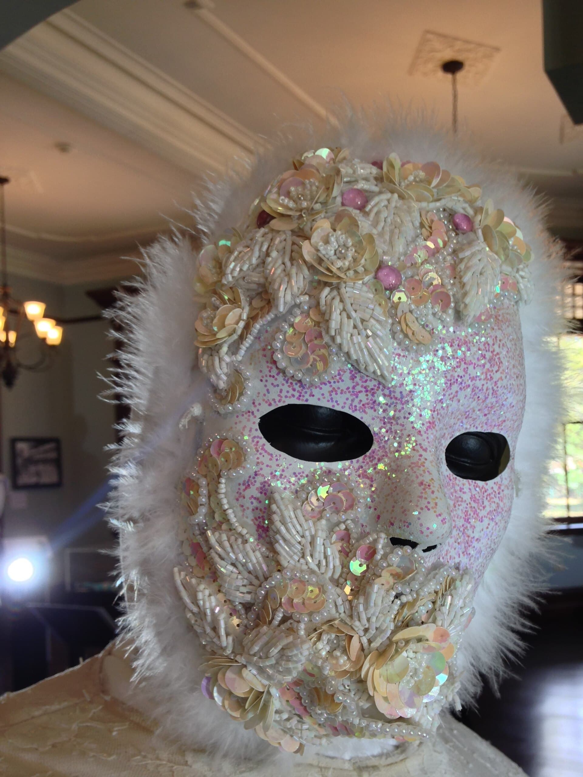 A white Pierrot mask by Peter Minshall covered in pearl beads, iridescent sequins and pink glitter, framed by white marabou feathers, on display indoors.