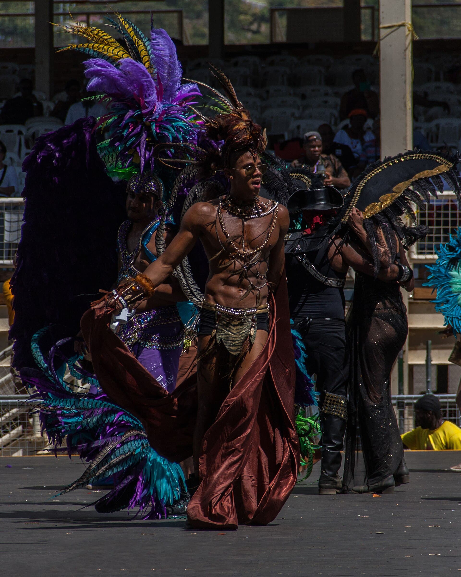 Masqueraders in towering purple, teal and black feathered costumes cross a stage during the Parade of the Bands at Trinidad Carnival 2015, with a man in a beaded harness and flowing bronze cape in front.