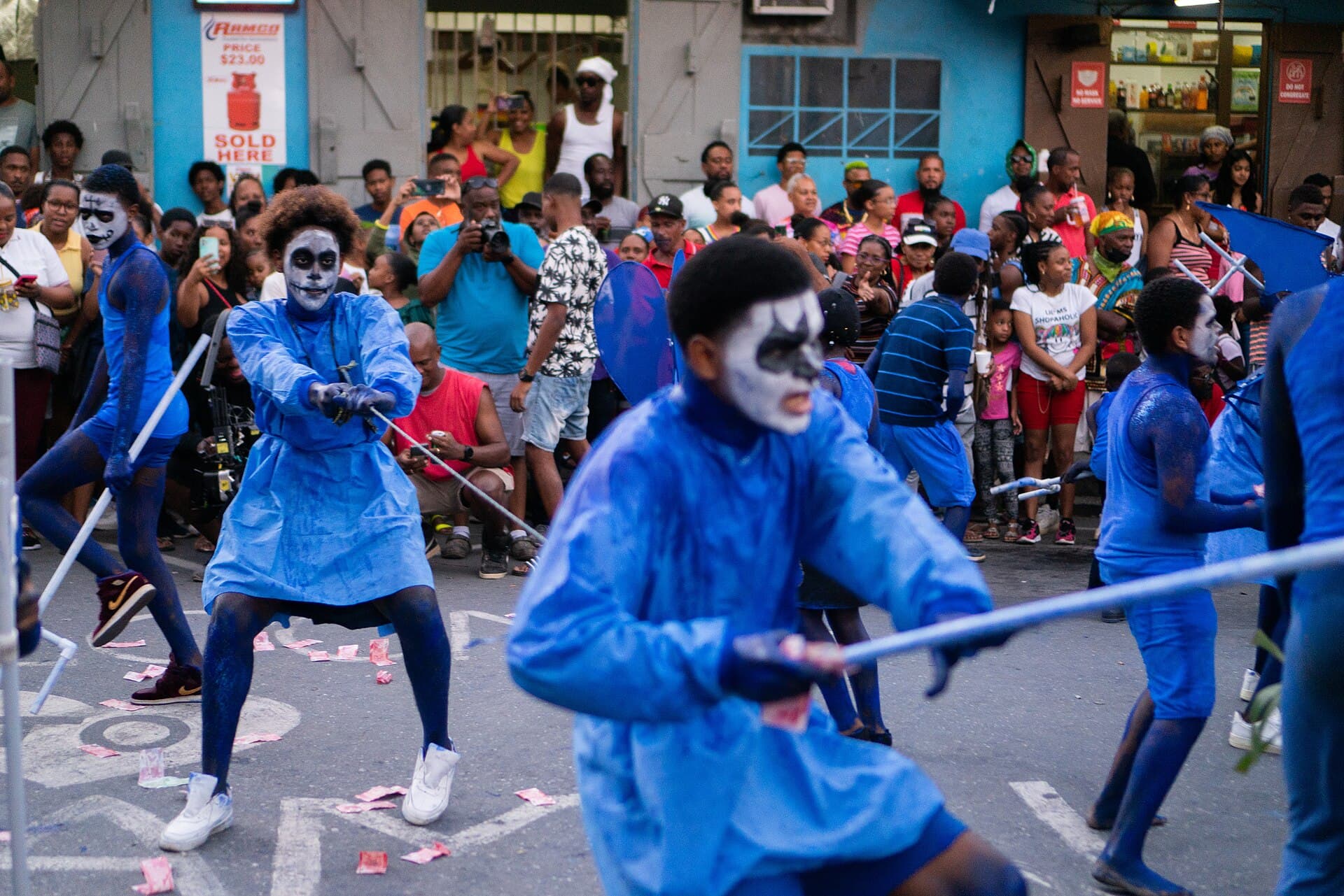 Young blue devil masqueraders at the Junior Jab competition in Paramin, Trinidad, in February 2023, dressed and daubed in blue with faces painted white like skulls, holding long poles in the street as a crowd of onlookers watches from the roadside