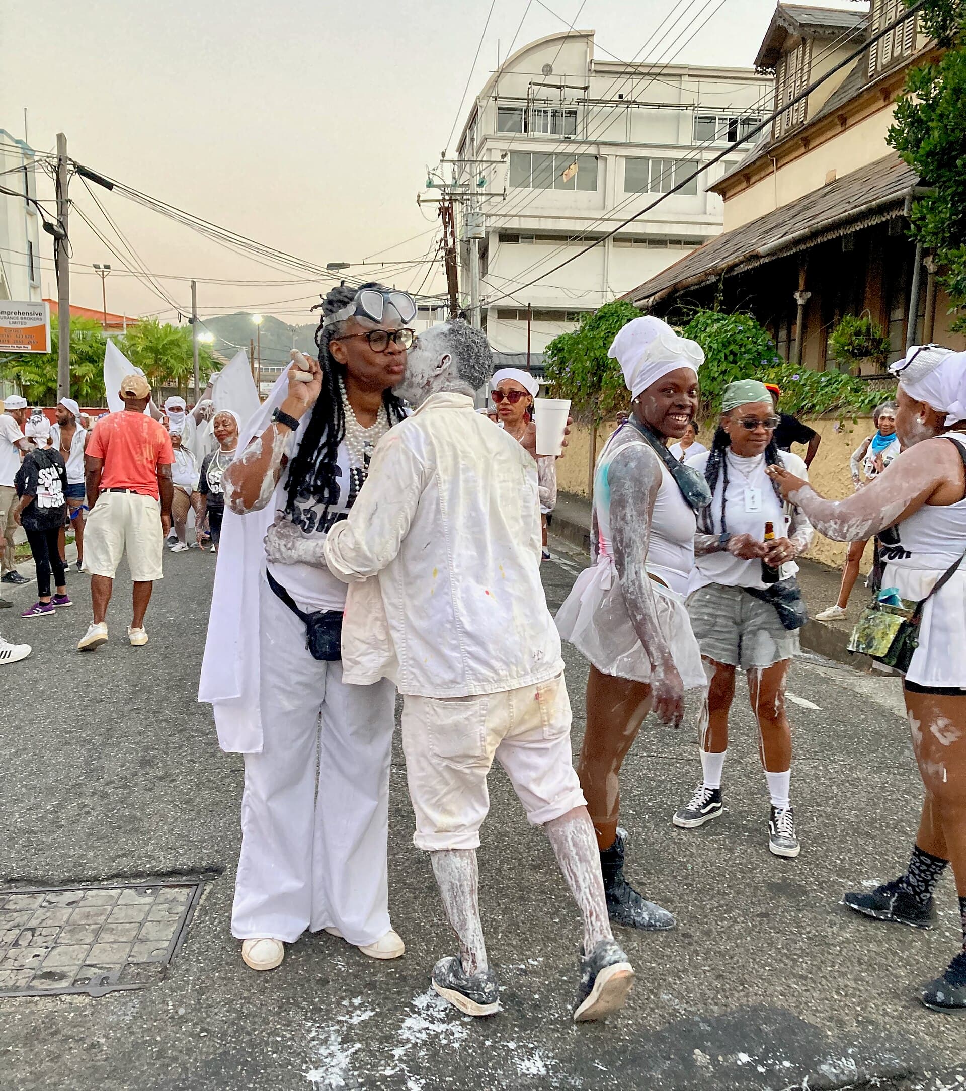 J'ouvert masqueraders from the 3Canal band, dressed in white and dusted with white powder and paint, greeting each other on a street in Port of Spain in early morning light at J'ouvert 2024, with more revellers behind them